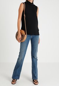 Sort ærmeløst top med høj hals, parret med lyseblå flared jeans. Holder en tan cirkulær taske med slangeskinds mønster.