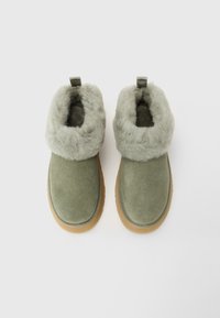 Pair of olive green suede slippers with thick, light gray faux fur lining and pull tabs, displayed on white background.