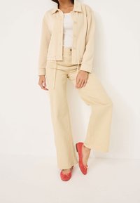 Chaqueta cropped beige con botones, parte superior blanca, pantalones beige de pierna ancha y bailarinas rojas con detalle de lazo. Tejido suave y corte relajado.