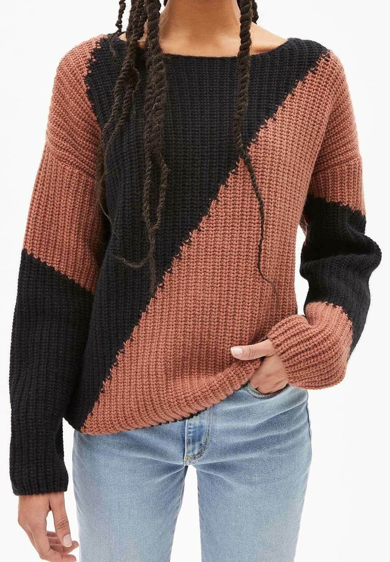 Personne portant un pull en maille côtelée avec des blocs de couleur diagonaux noir et marron, associé à un jean bleu clair, main dans la poche.