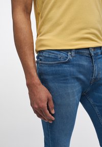 Siniset farkut, joissa on sileä tekstuuri, hieman haalistumista ja oranssia ommelta. Yhdistetty keltaiseen paitaan, joka ei ole kokonaan näkyvissä kuvassa.