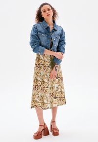 Jeansjacke mit knöpfbarer Vorderseite, getragen über einem bodenlangen Kleid mit Blumenmuster in Grün- und Brauntönen; das Modell trägt braune Plateauschuhe.