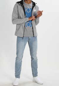 Grå zip-up hoodie med svarta detaljer, över en blå t-shirt med tryck, kombinerad med ljusblå jeans och vita sneakers.