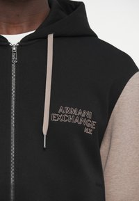 Sudadera con capucha negra y gris con cremallera, de textura suave. Presenta un logo bordado en gris claro en el pecho y una cremallera de contraste.