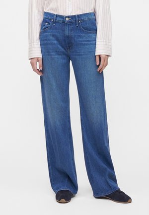 Personne portant un jean bleu taille haute à jambes larges avec une chemise à manches longues rayée rentrée à l'intérieur et des chaussures foncées sur fond blanc.