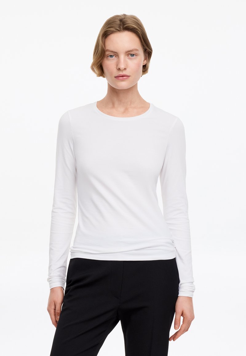 Femme aux cheveux courts châtain clair portant une chemise blanche unie à manches longues et un pantalon noir, regardant devant avec une expression neutre.