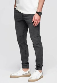 Jeans tapered gris oscuro con costuras laterales y puños de tobillo acanalados, combinados con zapatillas blancas y beige. Diseño simple sin patrones.