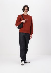 Roestrode sweatshirt, zwarte schoudertas, donkere jeans en witte sneakers met grijze accenten. Eenvoudig design, casual pasvorm, zachte textuur.