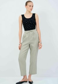 Černý top bez rukávů s bílými korálkovými akcenty, doplněný světlými vysokým pasem kalhot. Modelka má na nohou černé nazouvací boty.