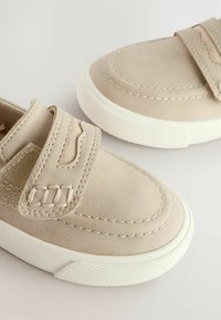 Beige mockaskor med vita gummisulor, med dubbel kardborreband och dekorativ söm längs ovandelen.