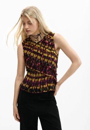 Kvinde med langt blondt hår iført en ærmeløs, flæset top med gult, pink og sort mønster, kombineret med sorte bukser, stående foran en hvid baggrund.