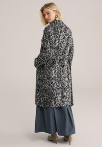Cappotto lungo con cintura in stampa animale grigia e nera, con una texture morbida. Il modello lo abbina a una gonna blu scuro e stivali taupe.