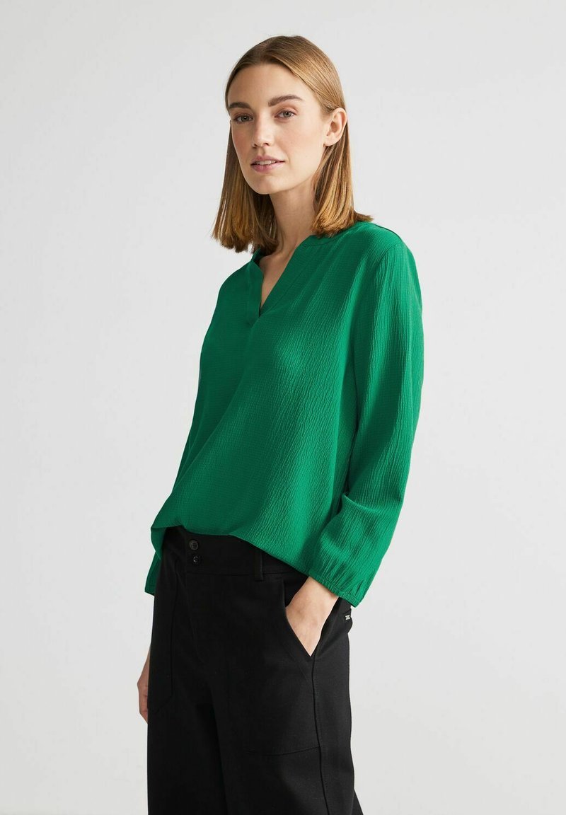Blusa verde, texturizzata, con scollo a V e maniche a tre quarti, abbinata a pantaloni neri. Caratterizzata da un design morbido e fluente.