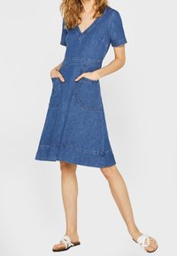 Denimmekko, jossa on lyhyet hihat, V-pääntie ja kaksi etutaskua. Vartalonmyötäinen yläosa ja levenevä hame, jossa on näkyviä ompeludetaileja.