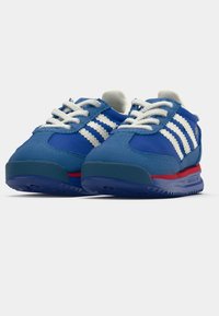 Zili sporta apavi ar baltām svītrām, ar teksturētu virsmu, noapaļotu purngalu un sarkanu akcentu gar zoli. Baltās šņorītes papildina dizainu.