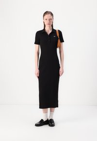 Juoda polo suknelė pritaikyto dizaino, trumpomis rankovėmis, siekia iki blauzdų. Šalia - šviesiai ruda petnešų rankinė ir juodi batai su baltais kojiniais.