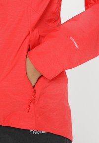 Veste rouge imperméable fabriquée en tissu texturé. Dotée d'une poche zippée et d'un logo "DRYVENT" sur la manche. Ourlet ajustable.