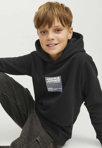 Felpa nera con cappuccio realizzata in tessuto morbido, caratterizzata da una toppa con logo sul davanti. Modello vestito con pantaloni grigio scuro, seduto davanti a uno sfondo neutro.
