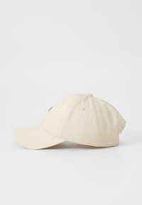 Beige baseballkeps med böjd skärm, textilstruktur och en broderad logotyp framtill. Har ventilationshål på sidorna.