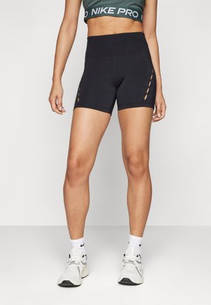 Vrouw draagt zwarte sportshorts met hoge taille en uitgesneden zijkanten, een groene Nike Pro sportbeha, witte Nike sokken en witte sneakers.