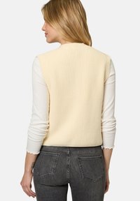 Rippstrick ärmelloser Pullover in blassgelb, getragen über einem weißen Langarmoberteil, kombiniert mit dunkelgrauen Jeans; schlichtes Design, weiche Textur.