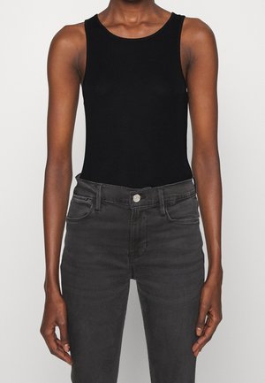Person iført en sort ærmeløs tanktop og højtaljede mørkegrå jeans, stående mod en ensfarvet lys baggrund.