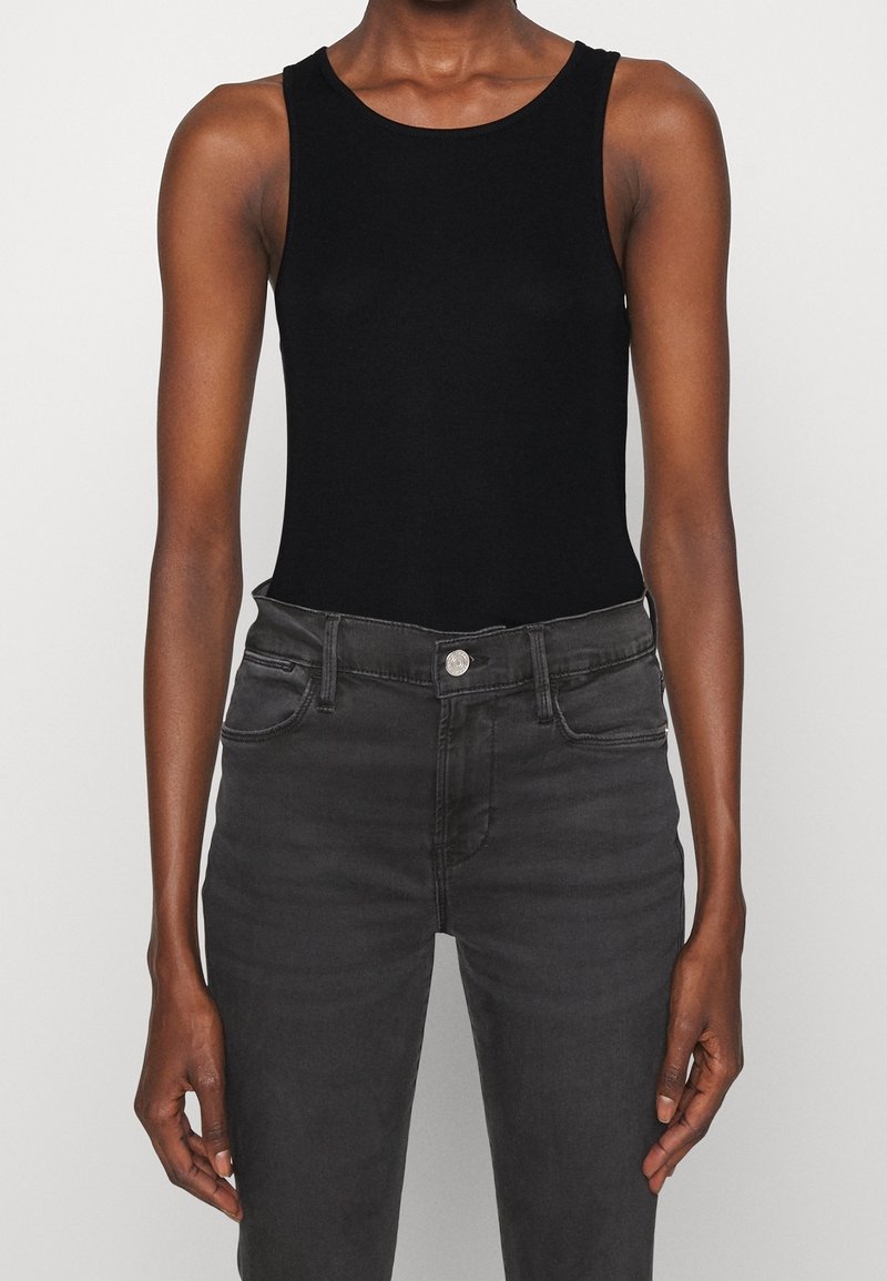 Personne portant un débardeur noir sans manches et un jean taille haute gris foncé, debout devant un fond clair uni.