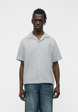 Junger Mann mit dunkler Hautfarbe und kurzen Dreadlocks, der ein hellgraues kurzärmliges Hemd mit Knöpfen und blaue Jeans trägt, vor einem weißen Hintergrund stehend.