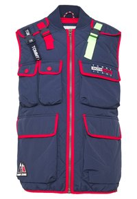 Navy quiltad väst med röda detaljer, framdragkedja, flera fickor och en logotypapplikation. Har justerbara remmar i rött och grönt.