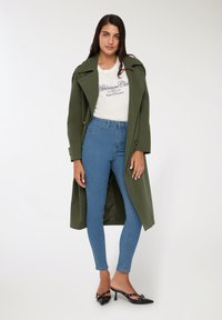 Oliwkowy trencz z dużymi klapami, noszony na białej koszulce z grafiką i niebieskich jeansach z wysokim stanem, w połączeniu z czarnymi, na obcasie klapkami.