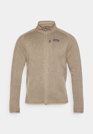 Veste en polaire couleur beige avec un col haut et une fermeture éclair sur toute la longueur. Présente des finitions beiges et un petit logo sur la poitrine gauche. Tissu texturé.