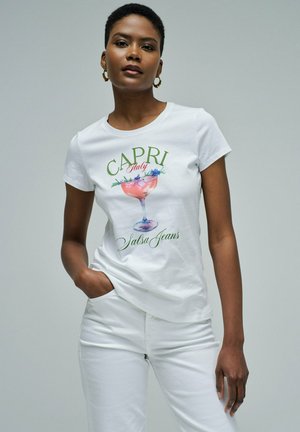 Mujer vistiendo una camiseta blanca con un vaso de cóctel rosa y texto verde "Capri Italy Salsa Jeans", combinada con pantalones blancos, posando contra un fondo gris.