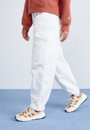 Pantalones cargo blancos con grandes bolsillos laterales y cierre de botón, combinados con zapatillas gruesas blancas y naranjas, sobre un suelo azul.
