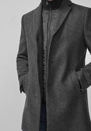 Mężczyzna w ciemnoszarym, teksturowanym płaszczu noszonym na pikowanej kurtce z zamkiem, ręce w kieszeniach, na tle jednolitego jasnego tła.