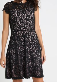 Schwarzes Spitzenkleid mit tailliertem Oberteil und ausgestelltem Rock, mit Blumenmustern und einer gewellten Ausschnittkante. Strukturierter Spitzenüberzug.