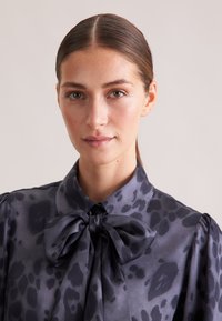 Blusa a macchie nere e grigie realizzata in tessuto morbido, dotata di un colletto con un grande fiocco e maniche a sbuffo arricciate.