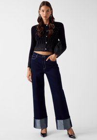Czarny krótki kardigan z srebrnymi ozdobami, zestawiony z granatowymi szerokimi spodniami jeansowymi z wysokim stanem, wykończonymi mankietami z denimu i kontrastującym przeszyciem.