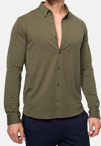 Olive green long-sleeve shirt with a button-down collar, light fabric, front placket with dark buttons, and a rounded hem.