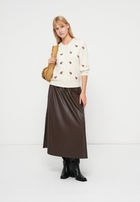 Cardigan crème avec broderies florales, associé à une jupe longue en simili cuir marron et des bottes noires montantes, tenant un sac à main beige.