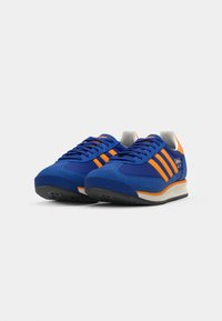 Blue suede sneakers with orange stripes, white accents, and a grey outsole. Features a padded collar and a textured rubber sole.