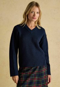 Marineblauer gestrickter Pullover mit V-Ausschnitt und langen Ärmeln, kombiniert mit einem karierten Rock in den Farben Grün, Rot und Gelb.