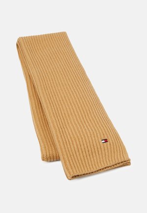 Écharpe en tricot côtelé beige pliée avec un petit logo drapeau rectangulaire rouge, blanc et bleu près d’un bord sur fond uni.