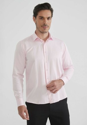 Homme aux cheveux bruns portant une chemise légère rose et un pantalon noir, ajustant la chemise au niveau de la taille sur un fond blanc.