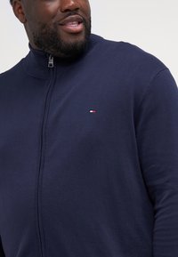 Bărbat purtând un pulover bleumarin cu fermoar și un mic logo alb și roșu pe piept, zâmbind ușor, pe un fundal simplu.