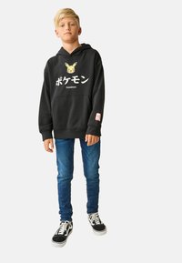 Black hooded sweatshirt with Pikachu graphic and Japanese characters; paired with blue jeans and black sneakers with white soles.