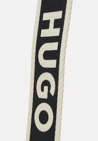 Schwarz-cremefarbener Riemen mit dem eingeprägten Schriftzug "HUGO" in einer fetten, modernen Schriftart, mit strukturierter Oberfläche und kontrastreichem Farbschema.