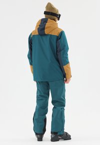 Teal waterproof jacket with tan accents, combined with matching pants. Features a hood, adjustable cuffs, and black lower pant sections.