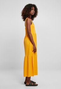 Geel, mouwloze maxi-jurk van zachte katoen met een gelaagde ruffle zoom; model draagt zwarte sandalen; eenvoudige, minimalistische stijl.