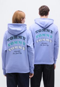Hellvioletter Kapuzenpullover mit großem, mehrfarbigem "TOMMY"-Schriftzug und dem Aufdruck "JEANS New York City" auf der Rückseite, hergestellt aus weichem Stoff.