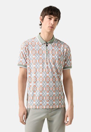 Νέος άνδρας που φορά κοντομάνικο πόλο με γεωμετρικό πορτοκαλί και γκρι σχέδιο και ανοιχτόχρωμο γκρι παντελόνι, στέκεται μπροστά σε απλό φόντο.